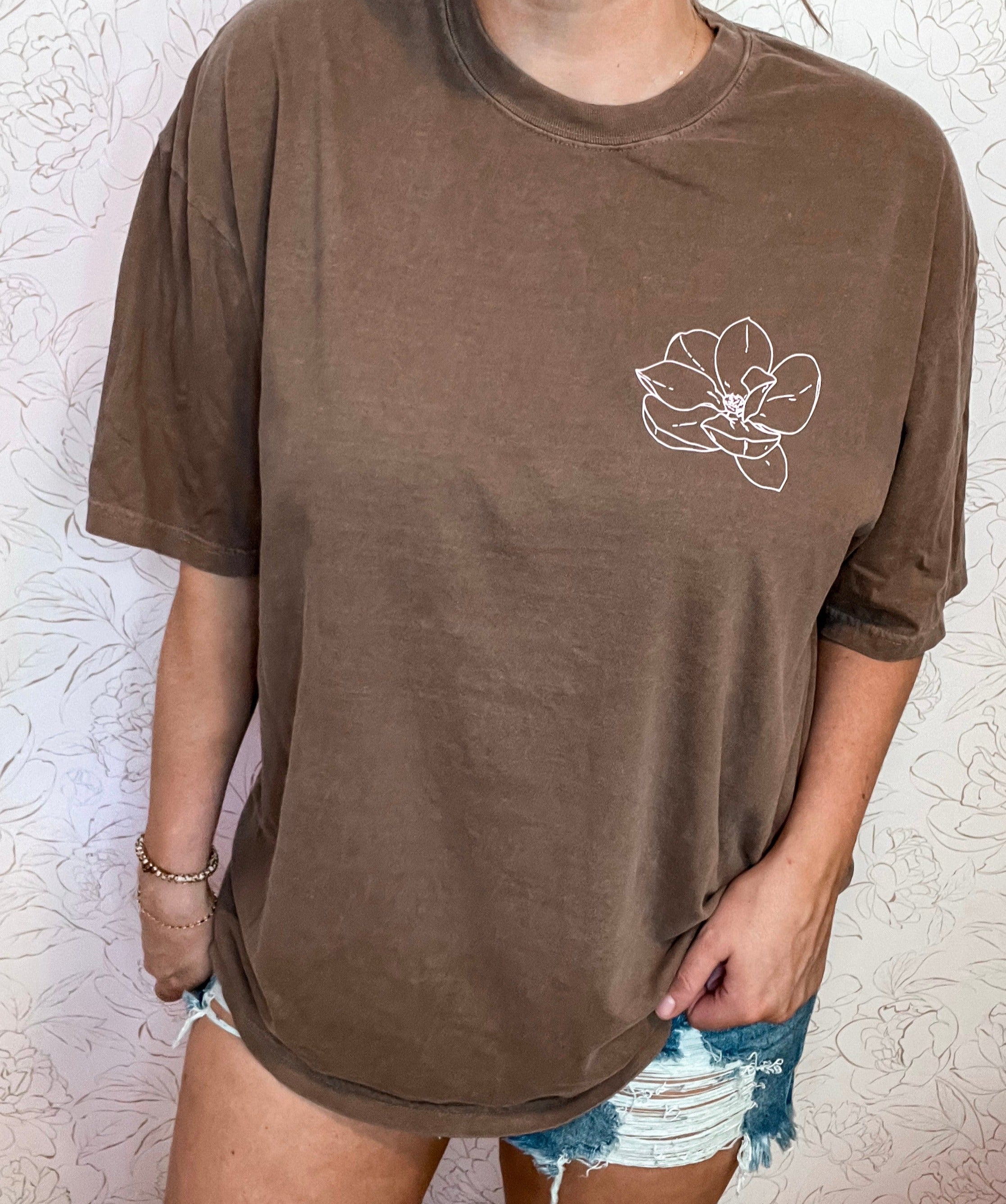 Stay stylishly comfortable with the Willow Grace and Co logo tee in rich espresso by Comfort Colors. This short-sleeved tee features a delicate pink magnolia flower in the top left corner, while the boutique's name is elegantly displayed in a soft baby pink on the back. Perfect for casual days or a touch of chic flair, this tee effortlessly combines comfort and charm