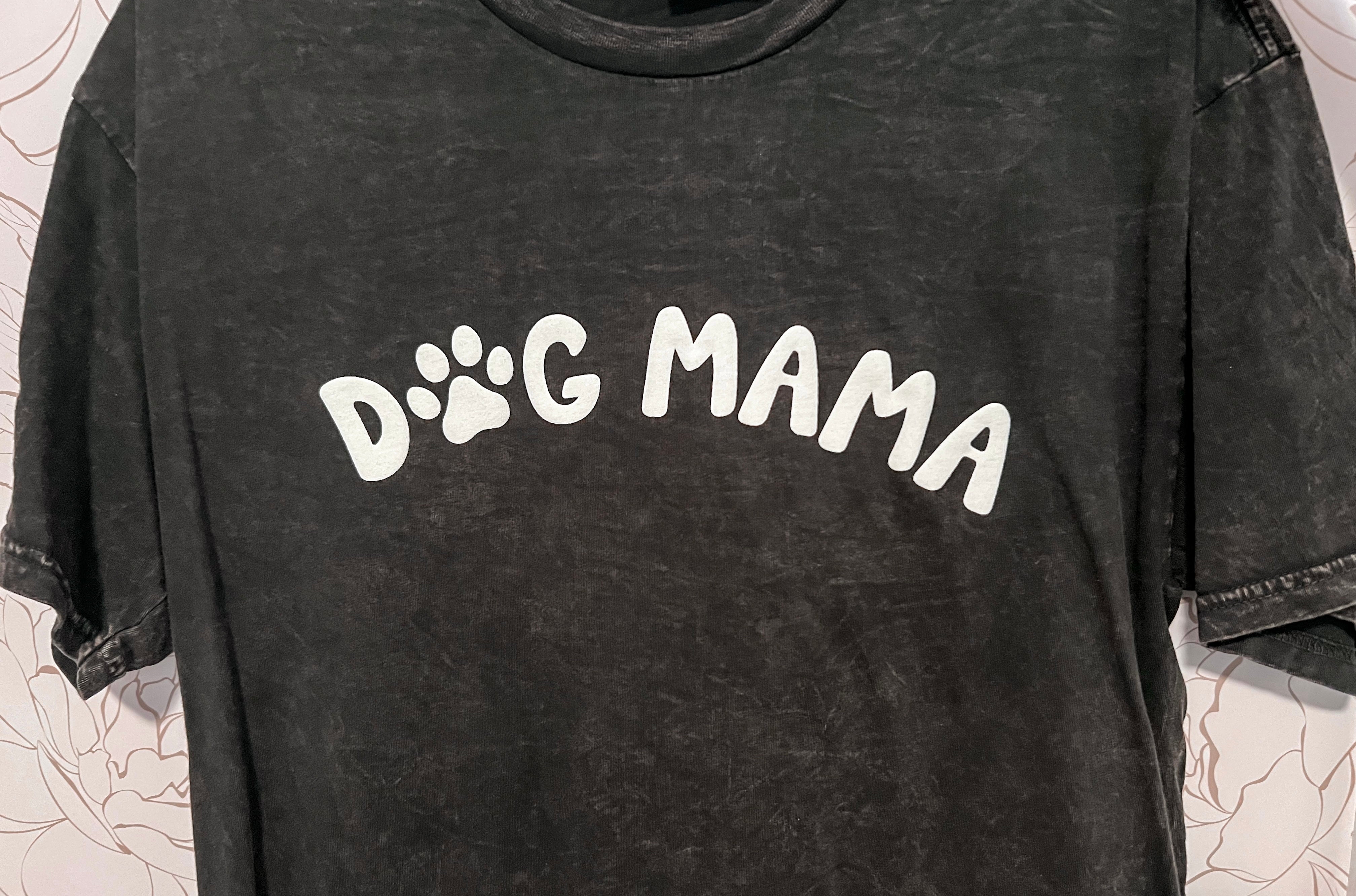 A black mineral-washed graphic tee with the phrase 'Dog Mama' prominently displayed in white puff print across the chest. The 'O' in 'Dog' is cleverly replaced with a white paw print, adding a playful touch to the design. The tee is crafted from a super soft and comfortable material, offering both style and comfort for dog lovers.