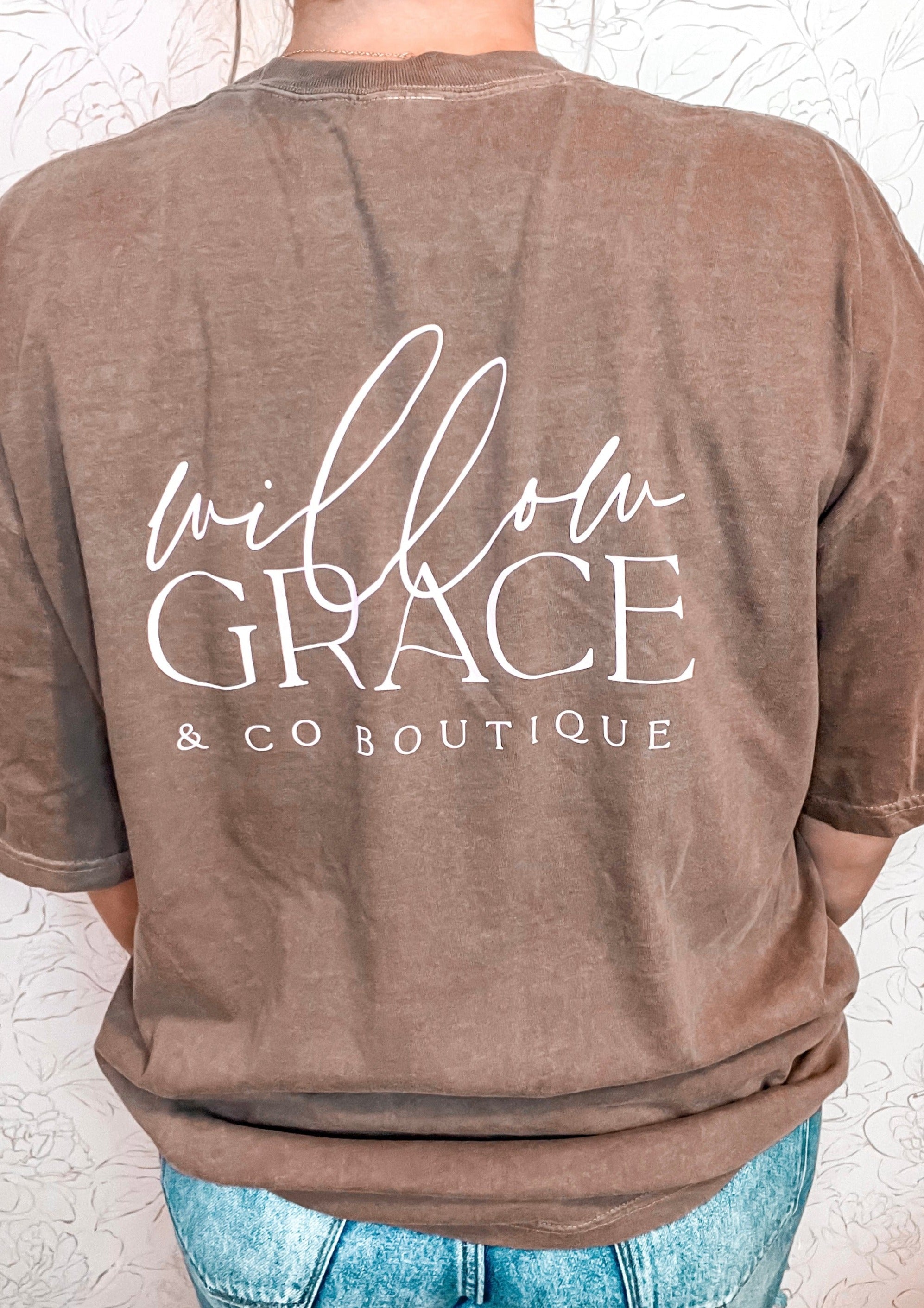 Stay stylishly comfortable with the Willow Grace and Co logo tee in rich espresso by Comfort Colors. This short-sleeved tee features a delicate pink magnolia flower in the top left corner, while the boutique's name is elegantly displayed in a soft baby pink on the back. Perfect for casual days or a touch of chic flair, this tee effortlessly combines comfort and charm