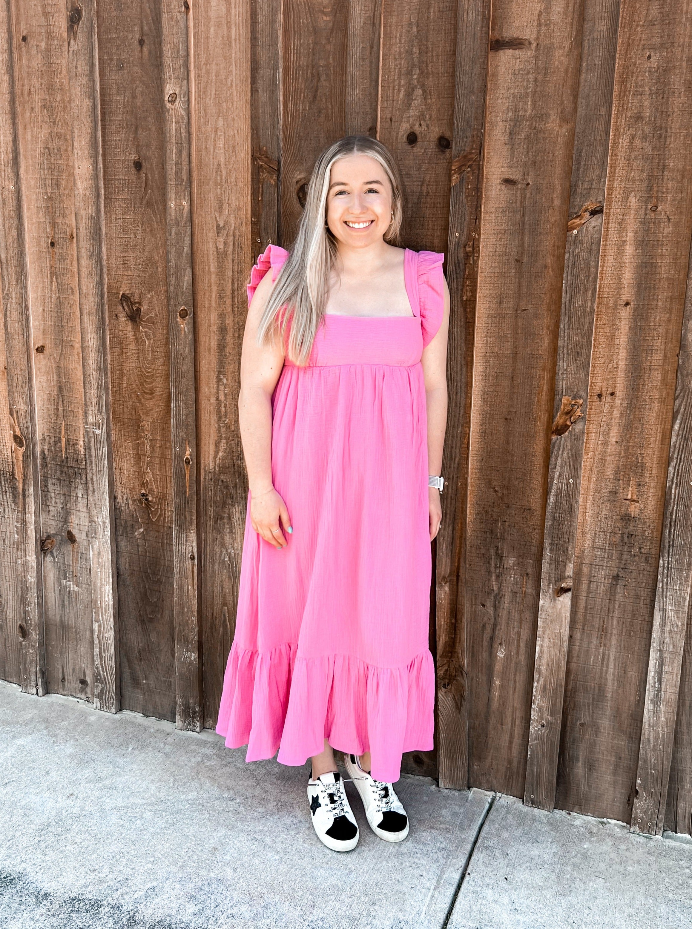 A bright pink maxi dress featuring ruffle cap/tank sleeves and an adjustable tie at the back, combining style with functionality. The dress has a tiered bottom that adds volume and movement to the silhouette, enhancing its playful and elegant appeal. This vibrant dress is perfect for warm-weather occasions, offering a blend of comfort and chic design.