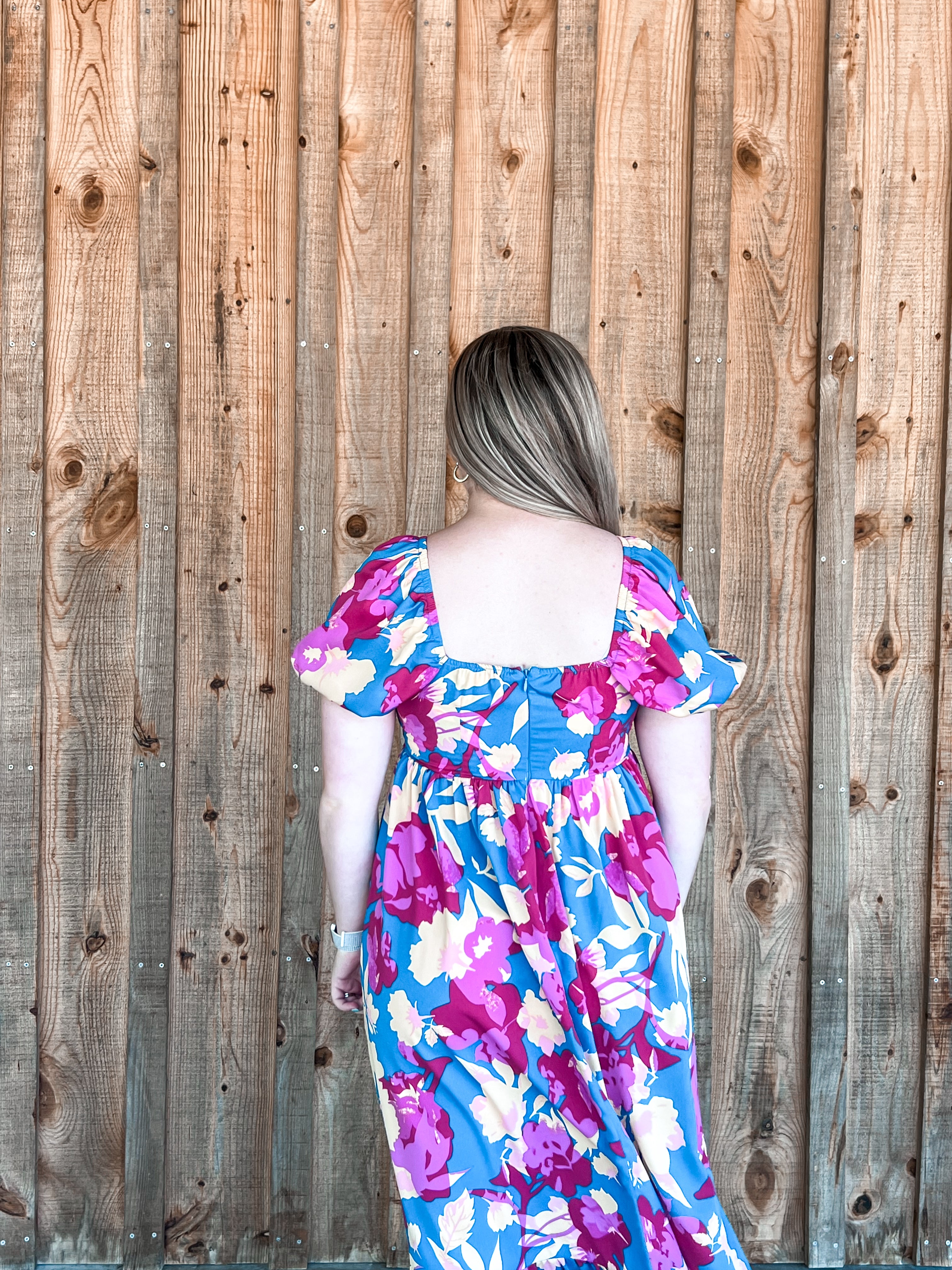 A boho-style maxi dress adorned with a floral pattern featuring vibrant colors of teal blue, purple, cream, and pink. The dress has short sleeves and a square neckline, enhancing its elegant and timeless appeal. Made from a cool material, this dress is suitable for all seasons, providing both style and comfort. Its vivid color palette and flowy silhouette encapsulate a festive and free-spirited vibe