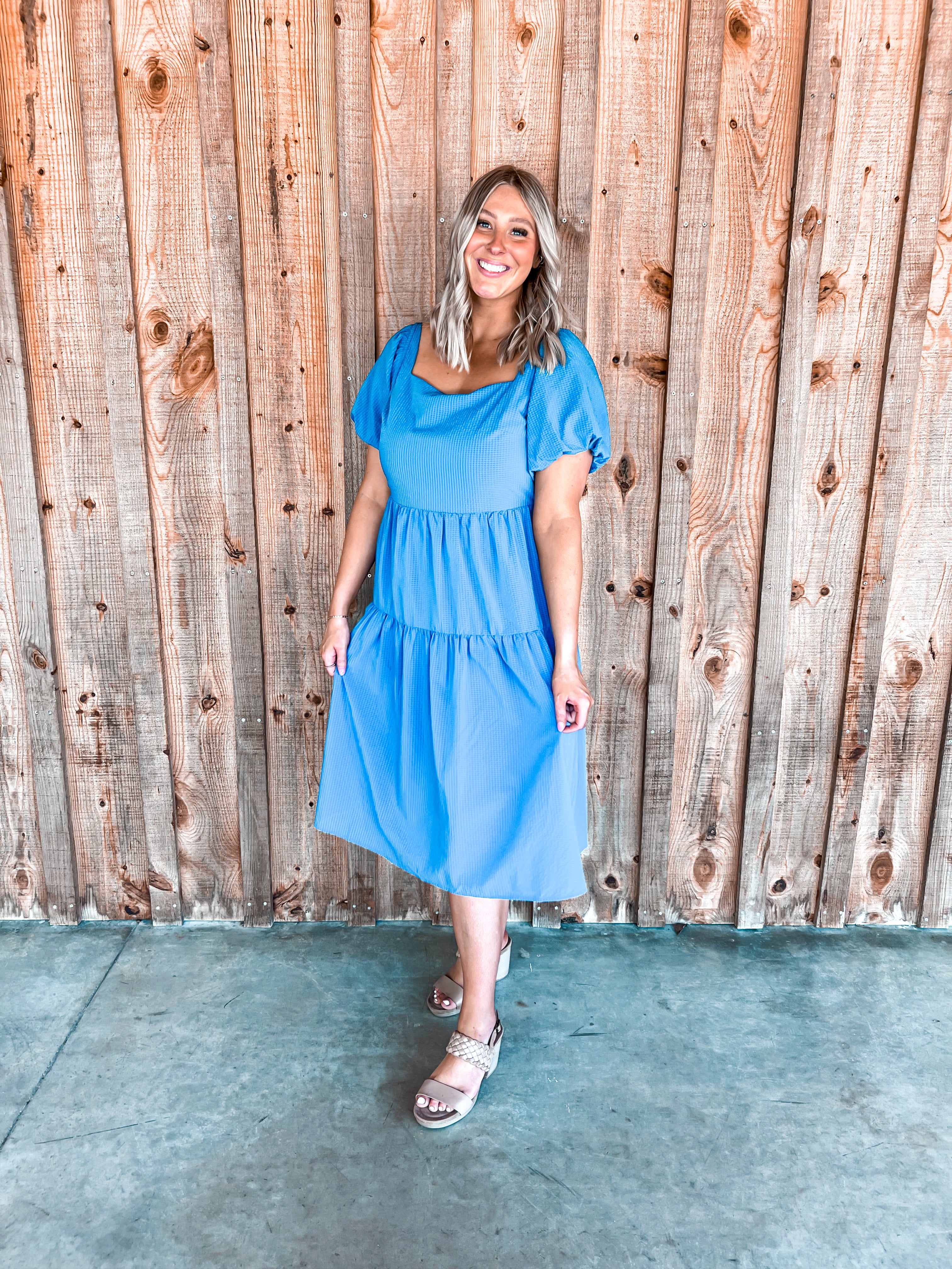 A cobalt blue midi-length dress with short puff sleeves and a charming tie detail at the back. It features a square neckline that flatters every figure and is made from a textured, cool material that adds a touch of intricate detail. The dress is designed with a tiered structure, enhancing its flowy silhouette and making it both stylish and comfortable for various occasions.