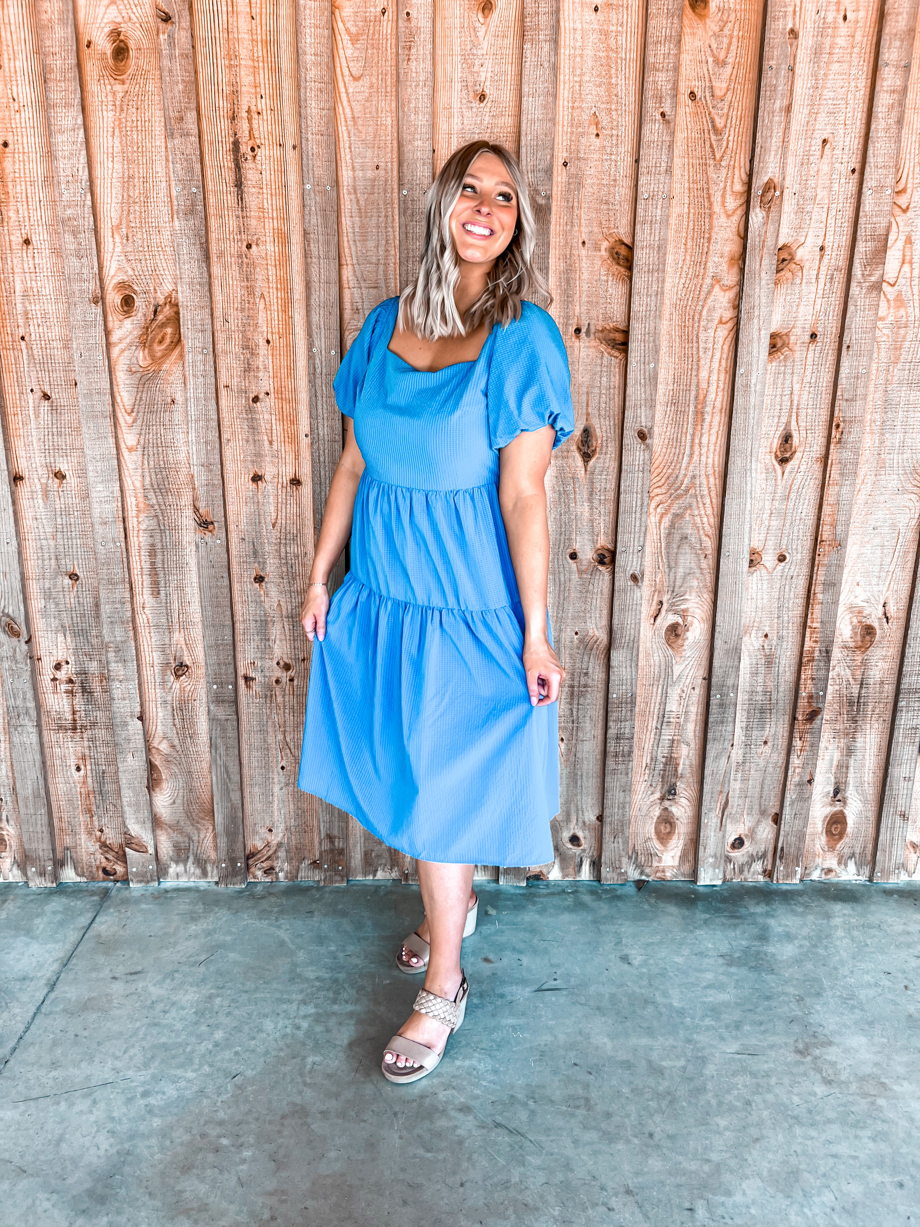 A cobalt blue midi-length dress with short puff sleeves and a charming tie detail at the back. It features a square neckline that flatters every figure and is made from a textured, cool material that adds a touch of intricate detail. The dress is designed with a tiered structure, enhancing its flowy silhouette and making it both stylish and comfortable for various occasions.