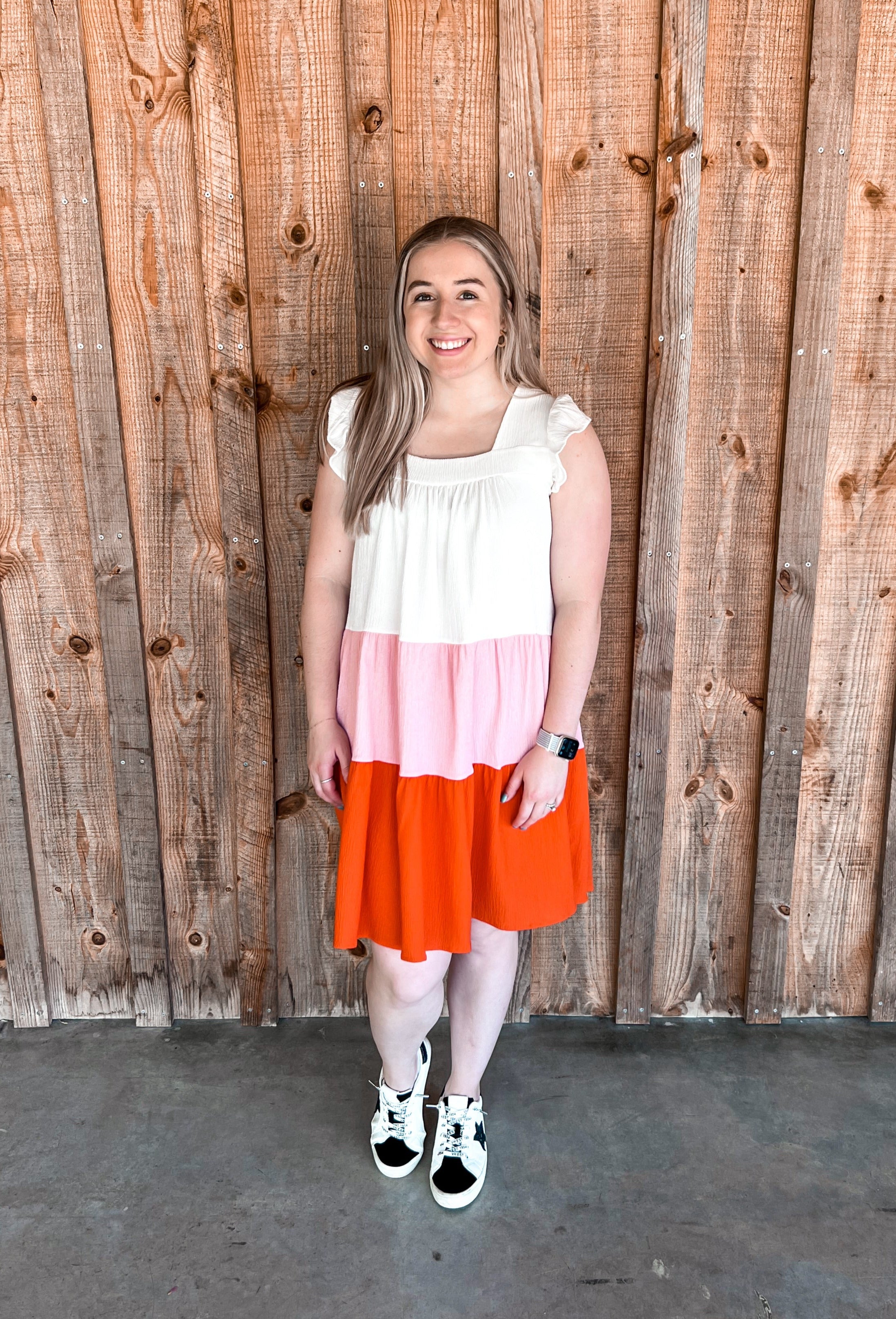 Above-the-knee, colorblock tiered dress featuring ruffle cap sleeves. The dress is designed with distinct horizontal sections of white, pink, and a reddish coral, creating a vibrant and summery feel. It is flowy, soft, cool, and comfortable, making it ideal for warm weather. The playful ruffles add a touch of femininity to the overall casual yet chic silhouette