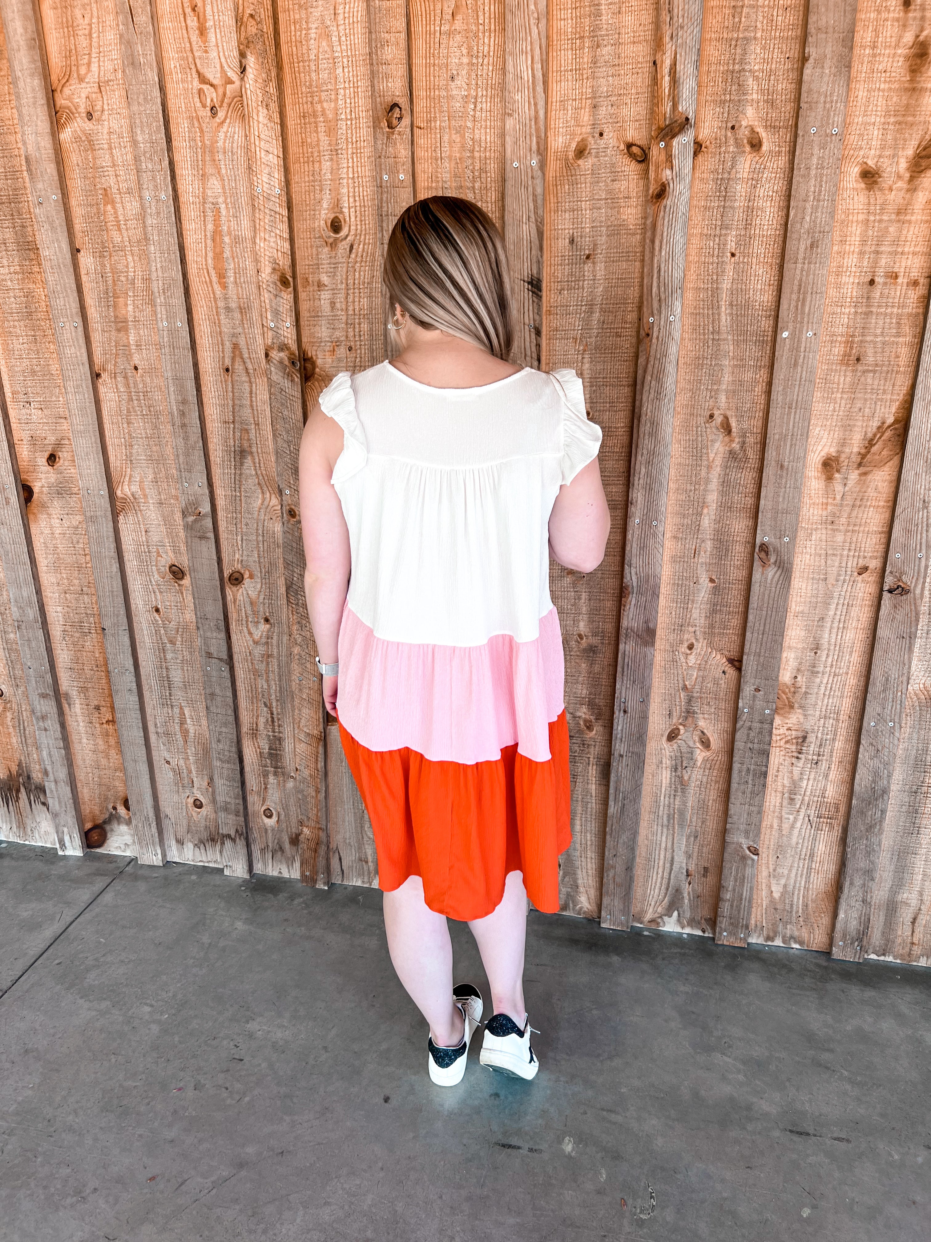 Above-the-knee, colorblock tiered dress featuring ruffle cap sleeves. The dress is designed with distinct horizontal sections of white, pink, and a reddish coral, creating a vibrant and summery feel. It is flowy, soft, cool, and comfortable, making it ideal for warm weather. The playful ruffles add a touch of femininity to the overall casual yet chic silhouette