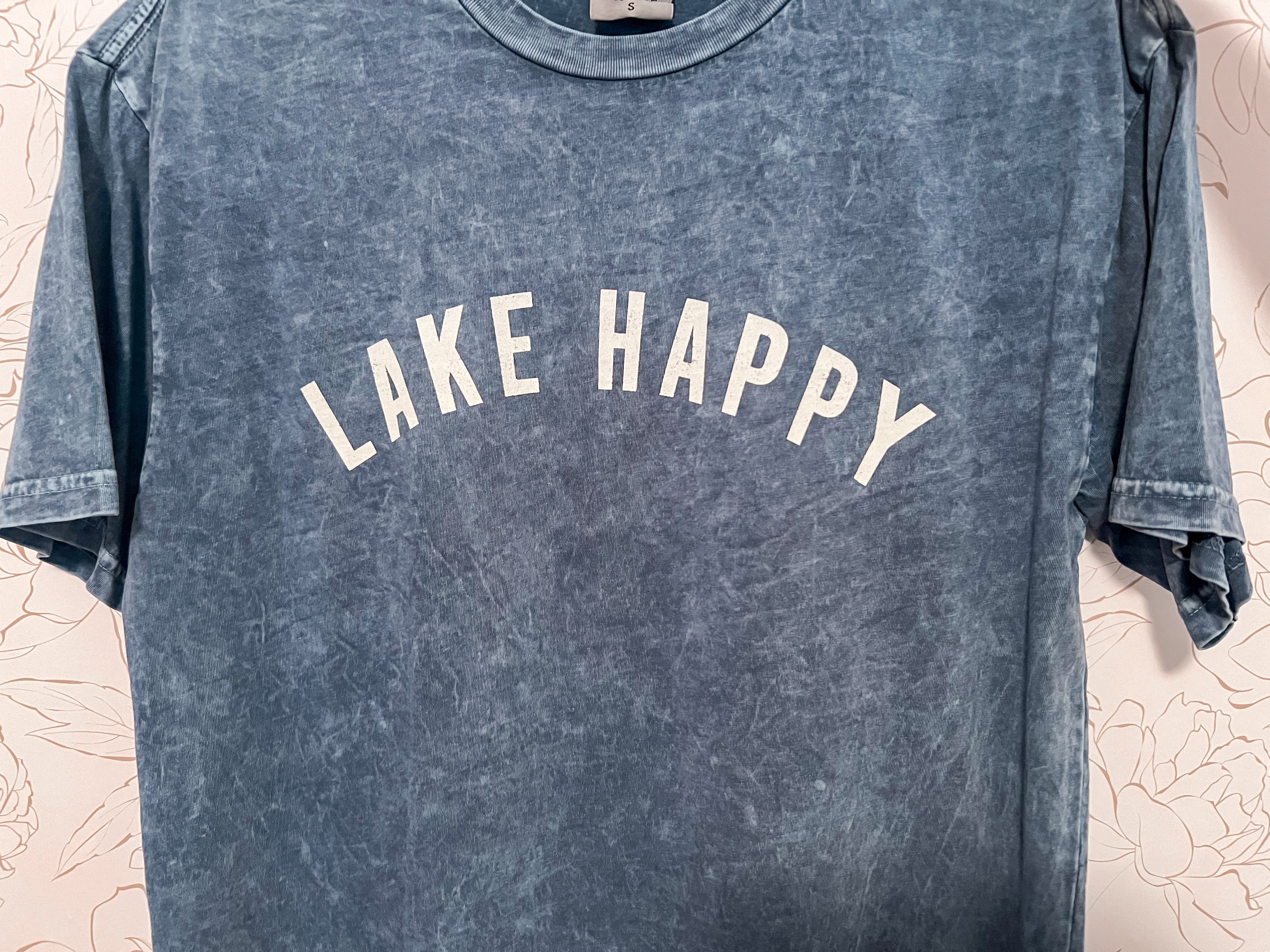 A mineral-washed blue graphic tee with the phrase 'Lake Happy' printed across the chest in white letters. The rich blue hue evokes the refreshing feel of summer by the water. The shirt's comfortable fit and inviting color make it an ideal choice for warm, laid-back days at the lake